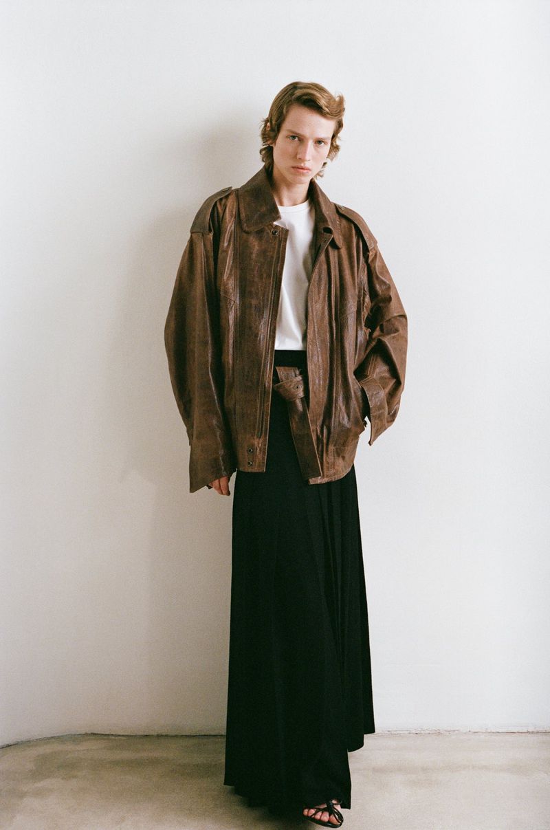 Fashion shot of a fair-skinned young adult in a brown leather jacket and long black skirt against an off-white studio backdrop.