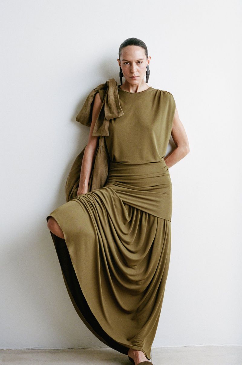 A stylish woman with dark hair is posing in a long olive-green draped dress and jacket against a white wall in a studio setting.