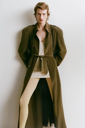 Model wearing an olive green belted trench coat over a cream top and pale tights against a plain studio background