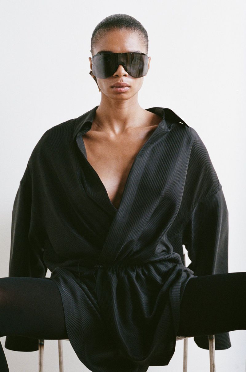 Woman with dark hair and sunglasses wearing a low-cut black shirt and black leggings sitting on a silver metal stool in a studio setting.