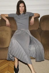 Model seated on a brown sofa wearing a grey knit top and a pleated grey skirt, for Erdos AW25.