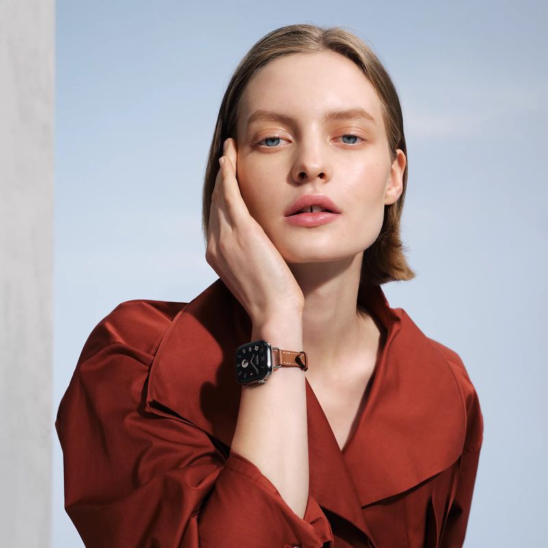 Portrait of fair-skinned woman with brown hair wearing a brown leather strap watch indoors against a soft blue and white background.