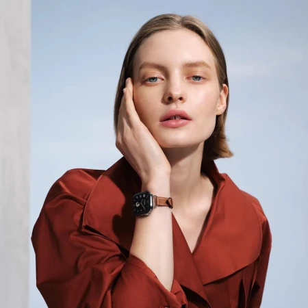 Portrait of fair-skinned woman with brown hair wearing a brown leather strap watch indoors against a soft blue and white background.