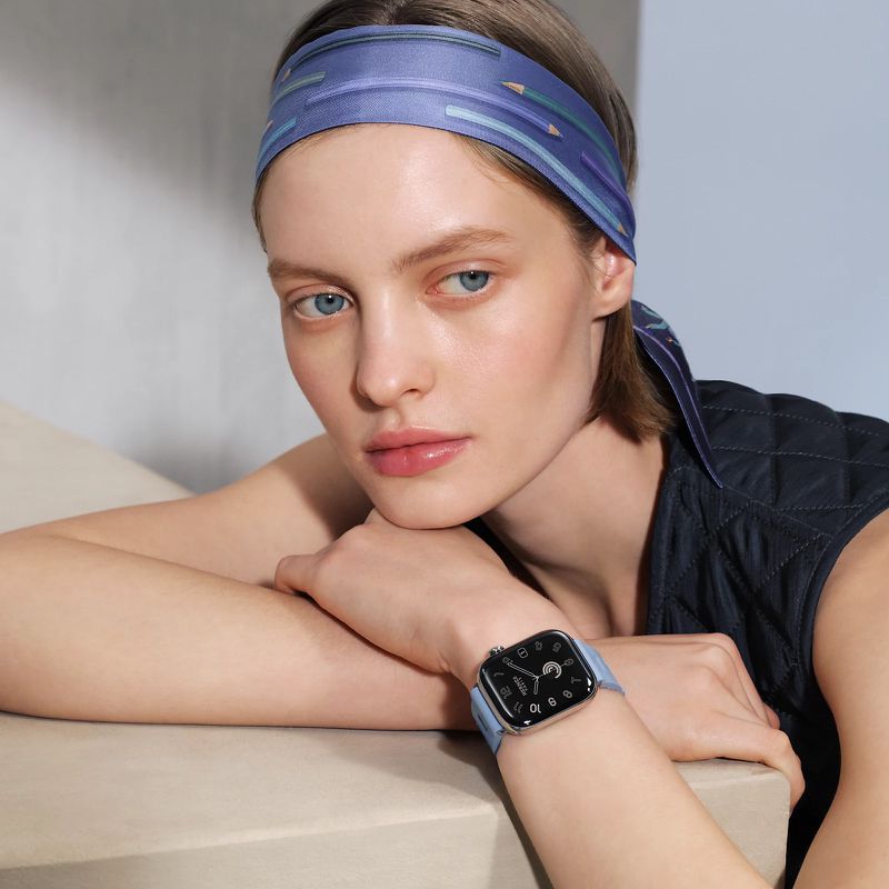 Close-up of a light-skinned woman wearing a patterned headscarf and smartwatch, resting her arms on a beige surface against a blue wall.