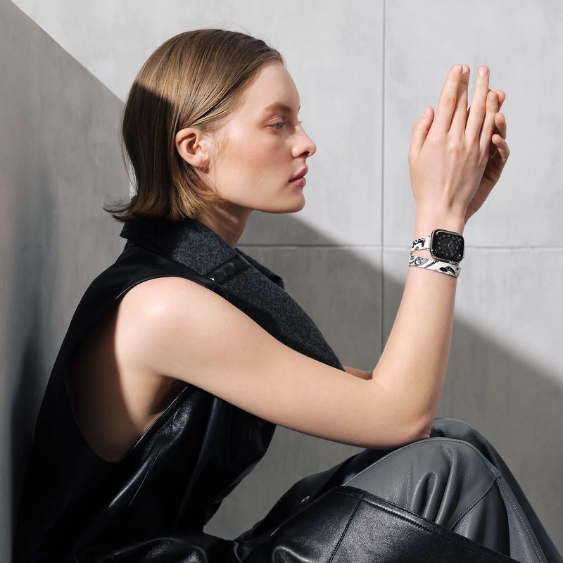 A woman with short blond hair wearing a black vest and holding up her hands to show off a patterned smart watch band.