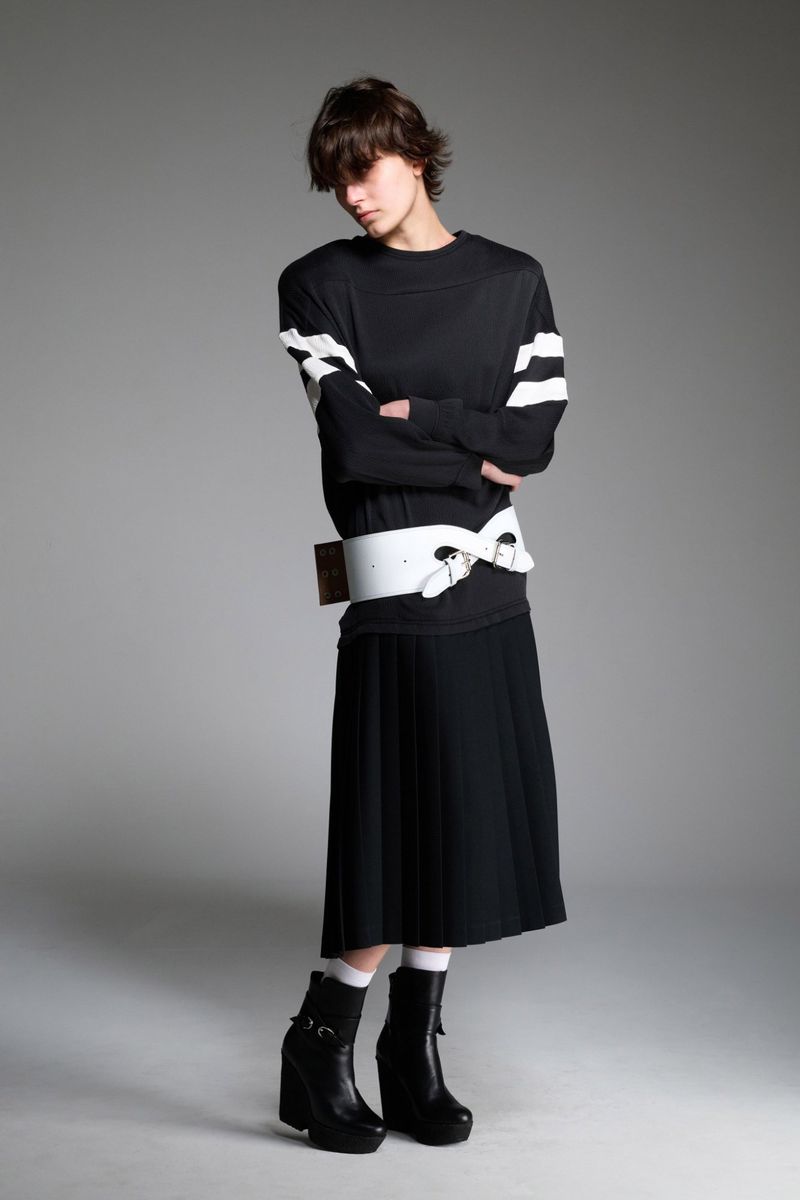 A person wearing a black, long-sleeved shirt with white stripes, a black skirt, and black boots standing in a photo studio.