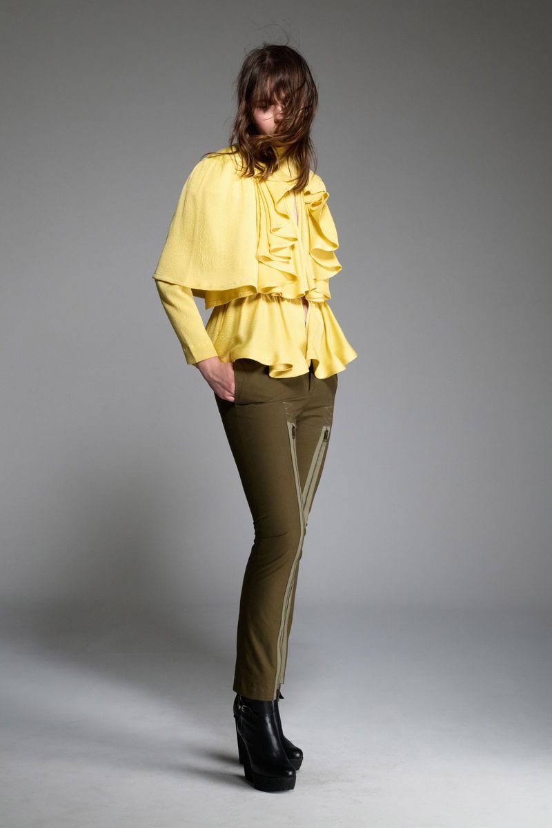 Studio shot of a woman in a yellow ruffled top, olive pants with silver zippers, and platform boots against a grey background.