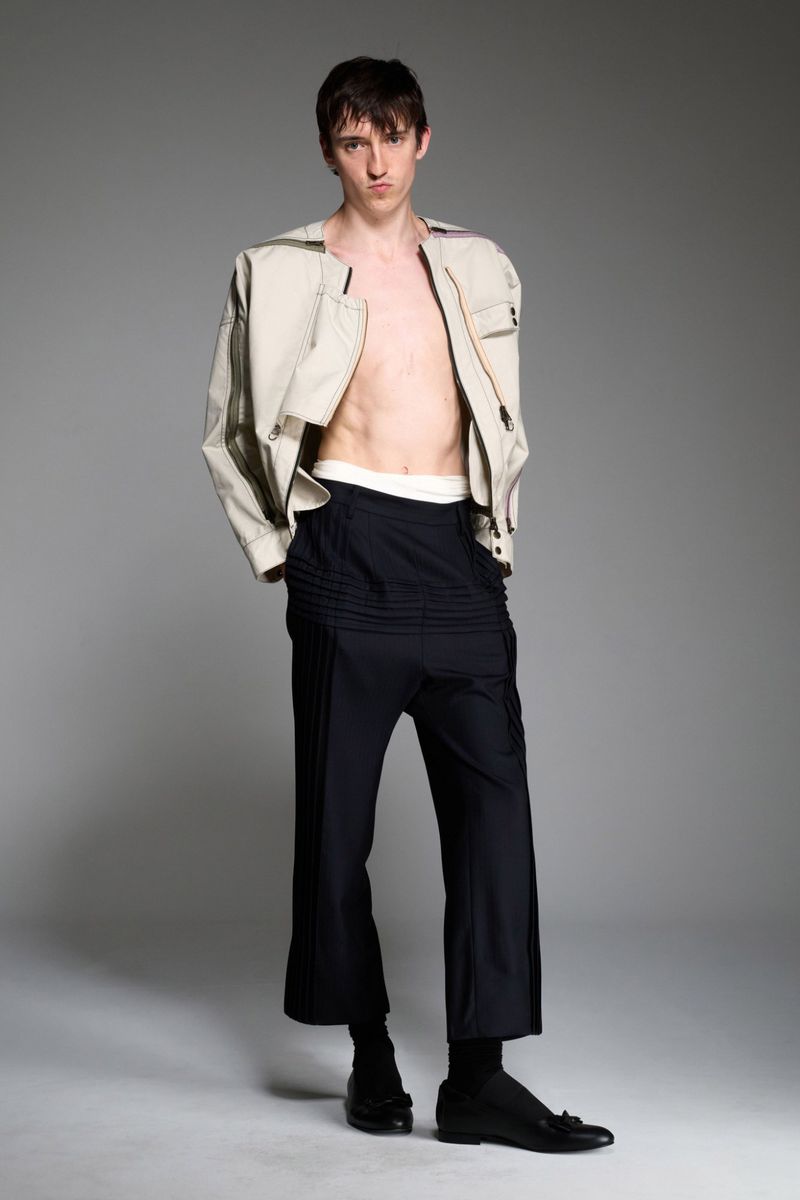 Full-length studio shot of a young adult male model wearing a beige jacket and navy cropped trousers against a neutral grey backdrop.