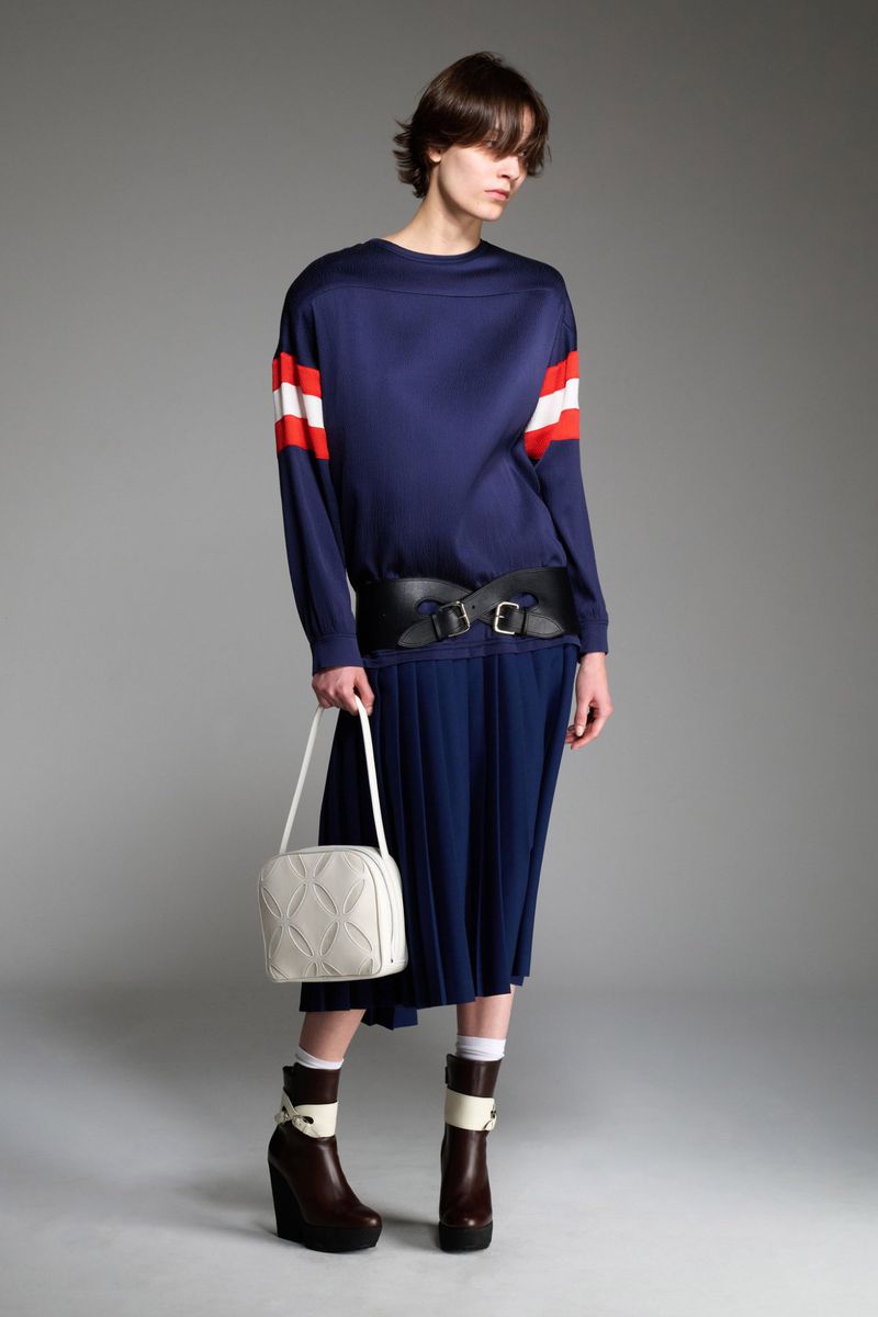 Fashion model poses in a navy long-sleeve shirt with red stripes, pleated skirt, wide black belt, white bag, and brown platform boots in studio.