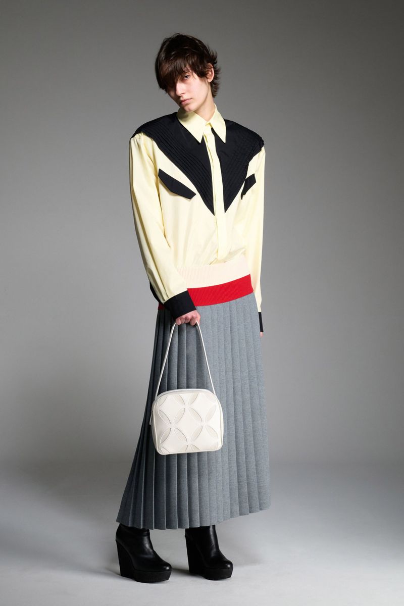 Adult female poses in a studio wearing a pale yellow blouse with black details, grey pleated skirt, and black platform shoes, holding a white bag.