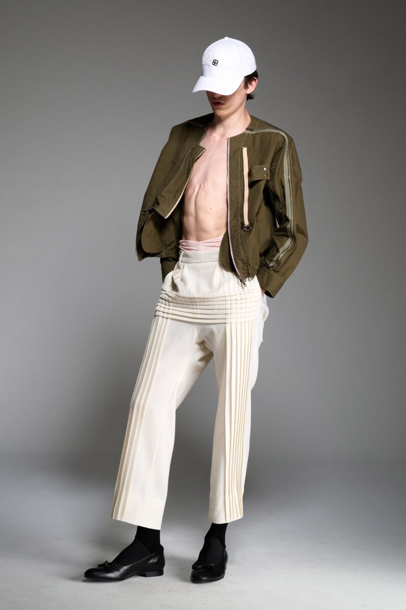 Full-length studio shot of an adult male wearing a baseball cap, open olive jacket, and cream trousers with pleats, posing against a gray background.