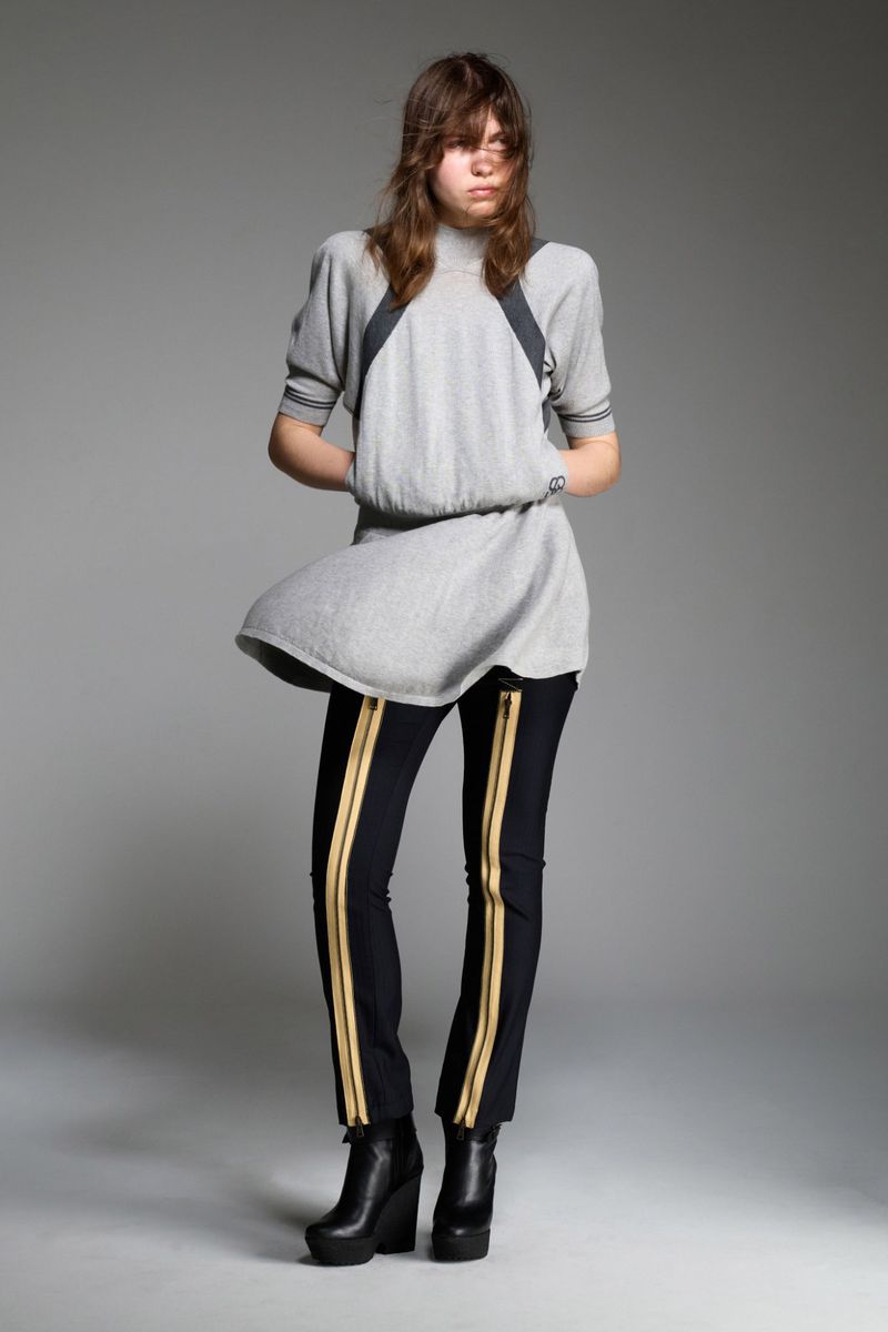 A young adult fashion model poses in a studio wearing a gray short-sleeve dress, black pants with gold stripes, and wedge platform boots.