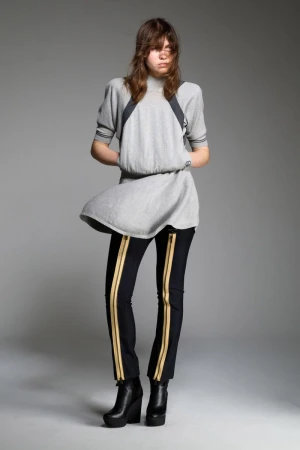A young adult fashion model poses in a studio wearing a gray short-sleeve dress, black pants with gold stripes, and wedge platform boots.