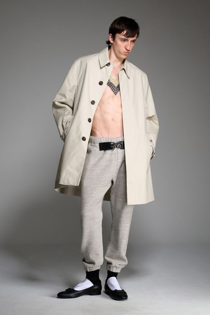 A slender adult male posing in a studio wearing a beige trench coat, grey sweatpants with a black belt, and black ballet shoes.
