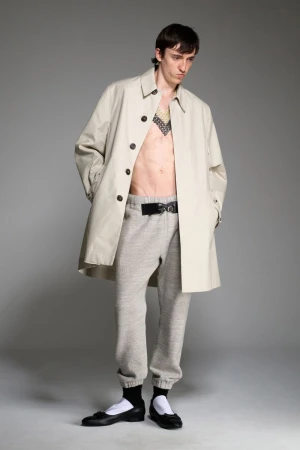 A slender adult male posing in a studio wearing a beige trench coat, grey sweatpants with a black belt, and black ballet shoes.