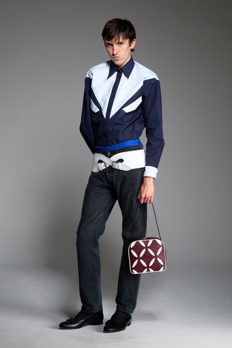 Fashion portrait of a young adult male in a patterned shirt and jeans holding a small purse in a photo studio with a neutral background.