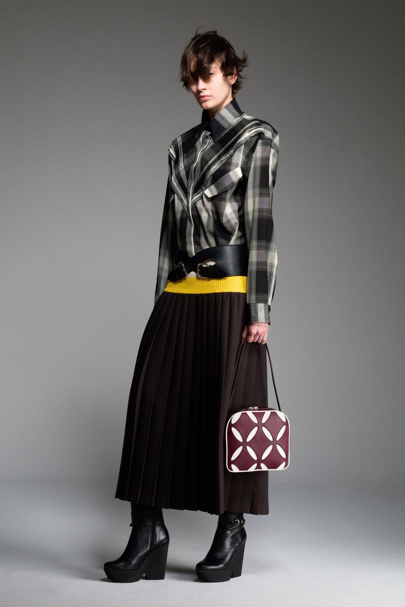 Fashion model in a studio setting wearing a plaid shirt, black skirt, yellow sweater, black belt, and black heeled boots, holding a purse.