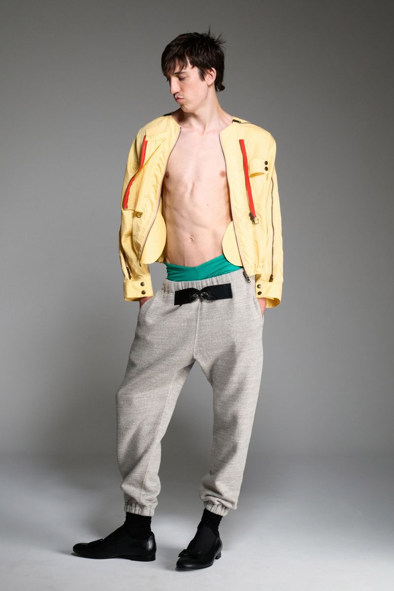 A slim, light-skinned adult male model stands in a studio wearing a yellow jacket, gray sweatpants, and black shoes in a fashion pose.
