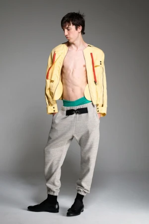 A slim, light-skinned adult male model stands in a studio wearing a yellow jacket, gray sweatpants, and black shoes in a fashion pose.