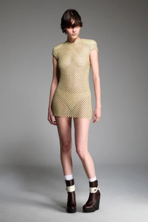 Fashion model posing in a short, woven, see-through dress with brown platform boots against a light gray studio backdrop.