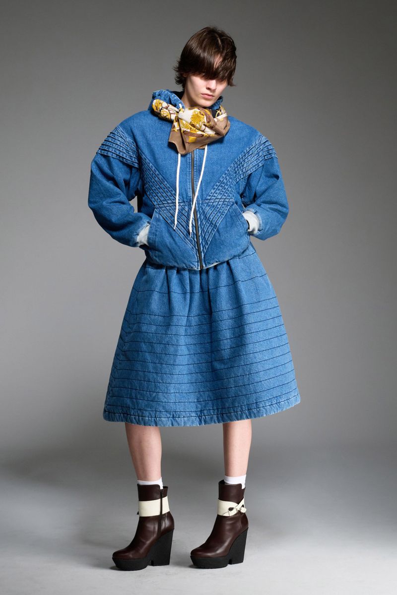 Fashion shot of a person in a blue denim jacket and skirt with brown wedge heel boots, standing in a photo studio against a grey backdrop.