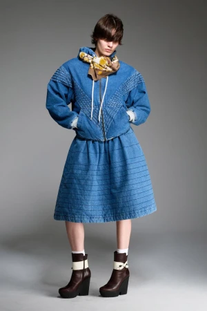 Fashion shot of a person in a blue denim jacket and skirt with brown wedge heel boots, standing in a photo studio against a grey backdrop.