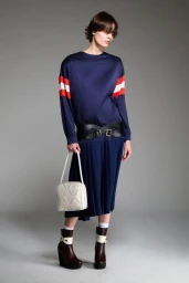 Model wearing a navy blue top with sleeve stripes, pleated skirt, and wide black belt holding a white bag against a grey backdrop.