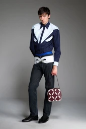Male model in a navy paneled shirt, wide belt, dark jeans, and holding a patterned burgundy handbag against a grey studio backdrop.
