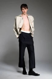 Male model in an open cream jacket and pleated black trousers posing against a neutral grey studio backdrop.