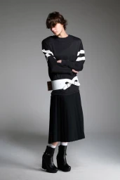 Model wearing a black striped top, wide white buckled belt, pleated midi skirt, and platform boots against a grey studio backdrop.