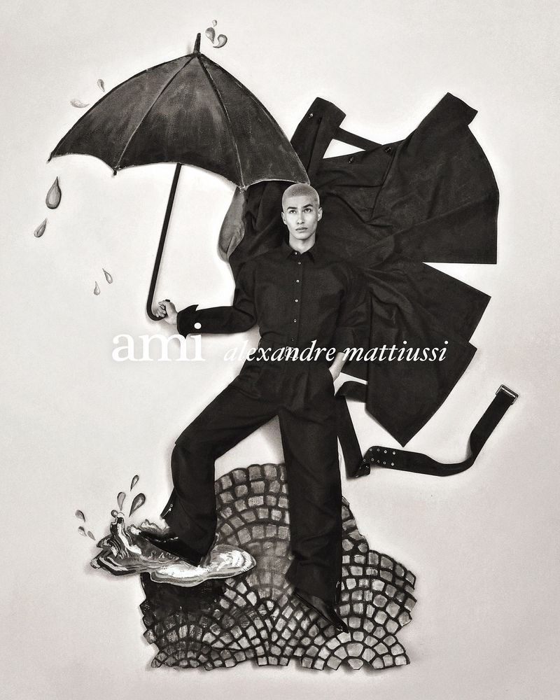 A male model in a black outfit holding an umbrella, standing on an elevated stone structure with a black coat in the background.