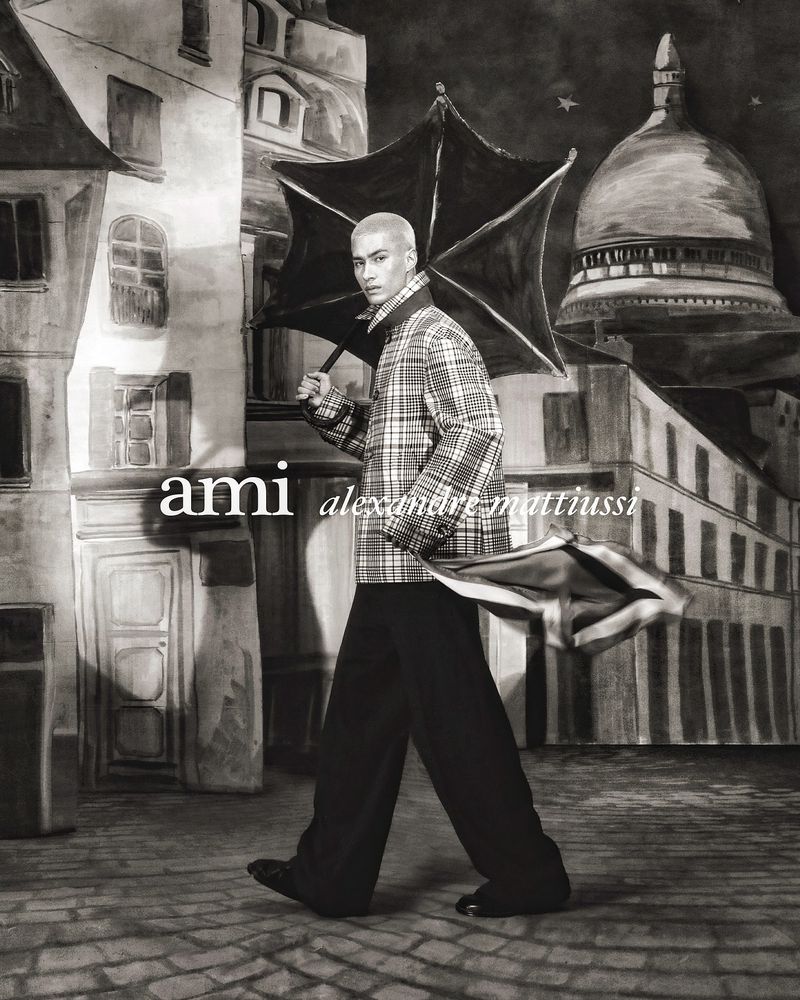 An adult male model poses with an umbrella in a faux painted-style backdrop resembling a city street, in a black-and-white artistic shot.