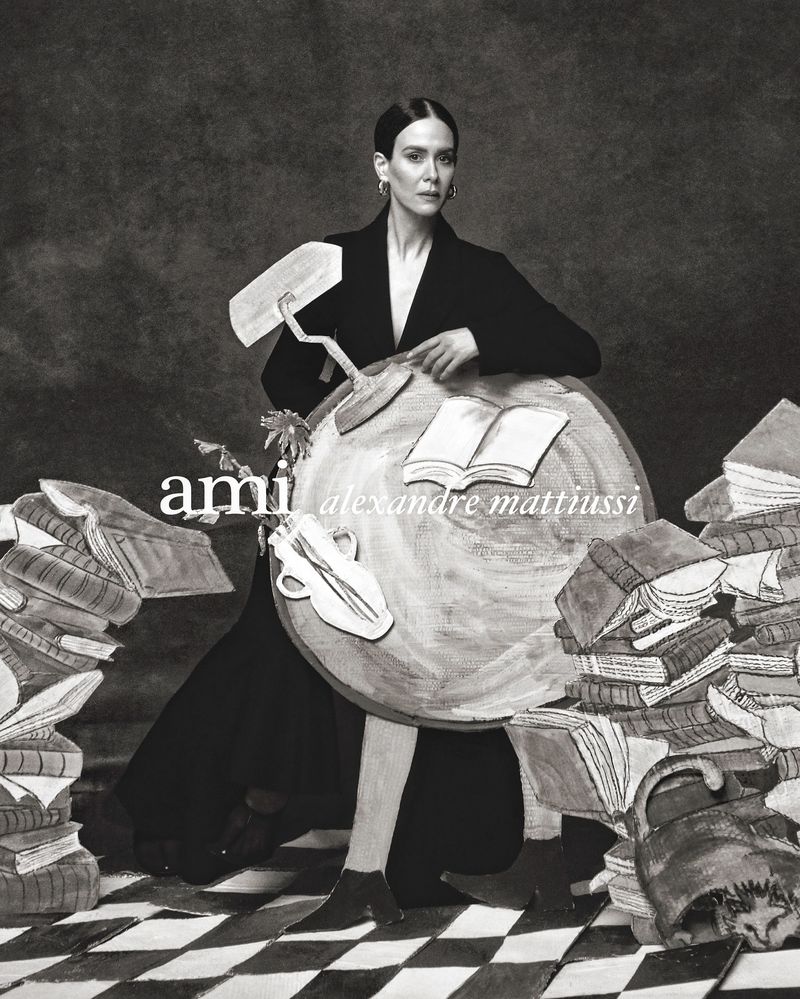 Adult female in a black coat poses in front of a black and white backdrop with painted books, accessories, and a checkerboard floor.