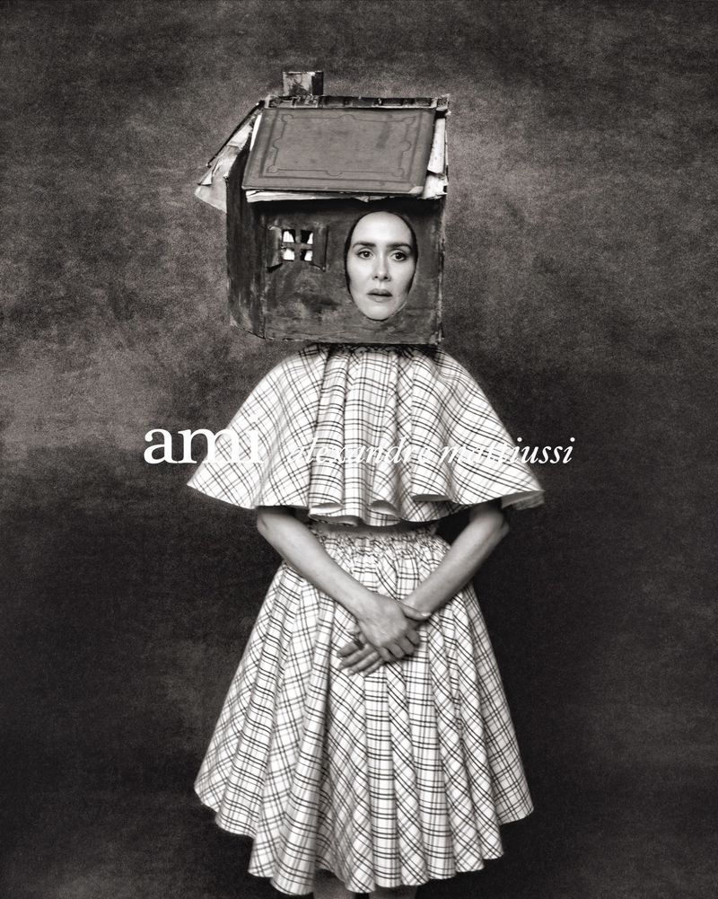 Monochrome portrait of a woman wearing a house-shaped headpiece and a plaid dress with hands clasped in front of her, against a mottled backdrop.