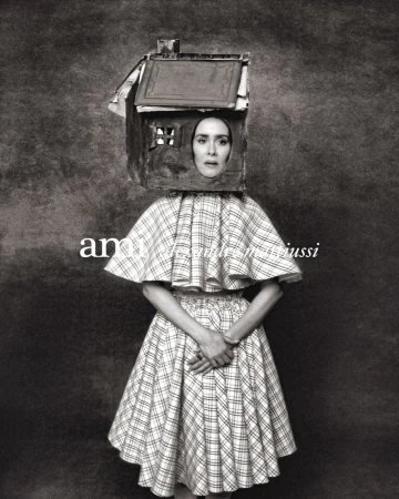 Monochrome portrait of a woman wearing a house-shaped headpiece and a plaid dress with hands clasped in front of her, against a mottled backdrop.