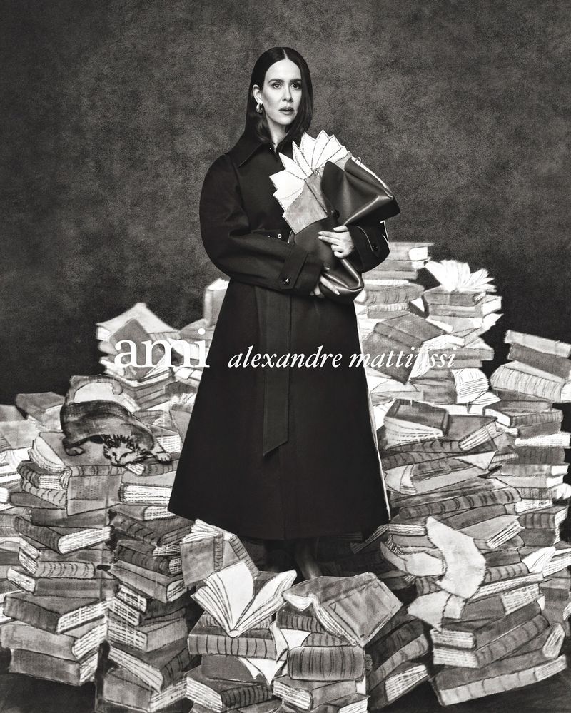 Sarah Paulson standing atop a large pile of books holding an open folder in an Ami Spring-Summer 2025 editorial photograph.