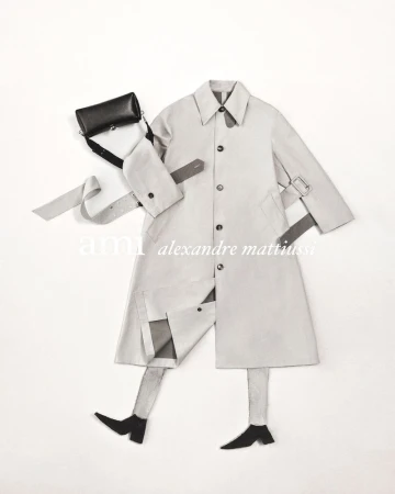 Fashion editorial featuring a beige trench coat laid flat on a white surface with a black cross-body bag and leg cutouts suggesting a person.