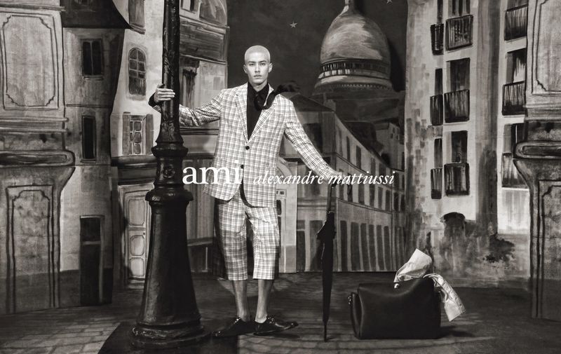 Man in plaid suit posing next to a lamp post with city painted backdrop, holding an umbrella and bag. Fashion editorial shoot.