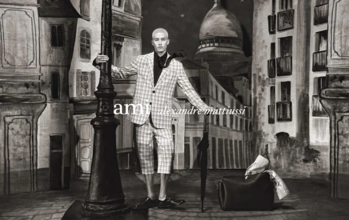 Man in plaid suit posing next to a lamp post with city painted backdrop, holding an umbrella and bag. Fashion editorial shoot.