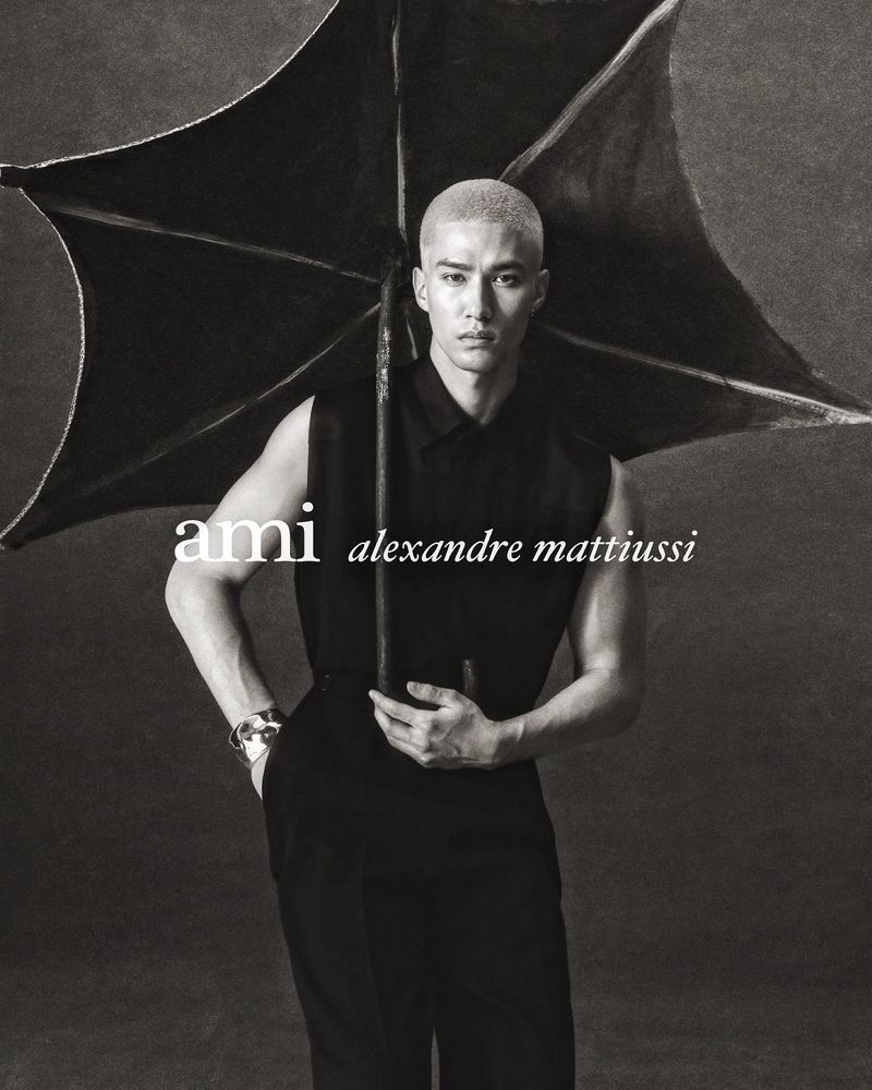 Monochrome fashion portrait of a man holding a black umbrella wearing a sleeveless black top against a dark textured background.