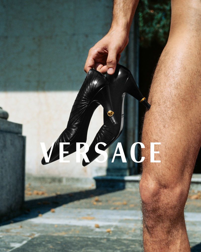 Versace fashion ad featuring a fair-skinned person holding a black leather high-heel shoe with gold medallion outdoors.