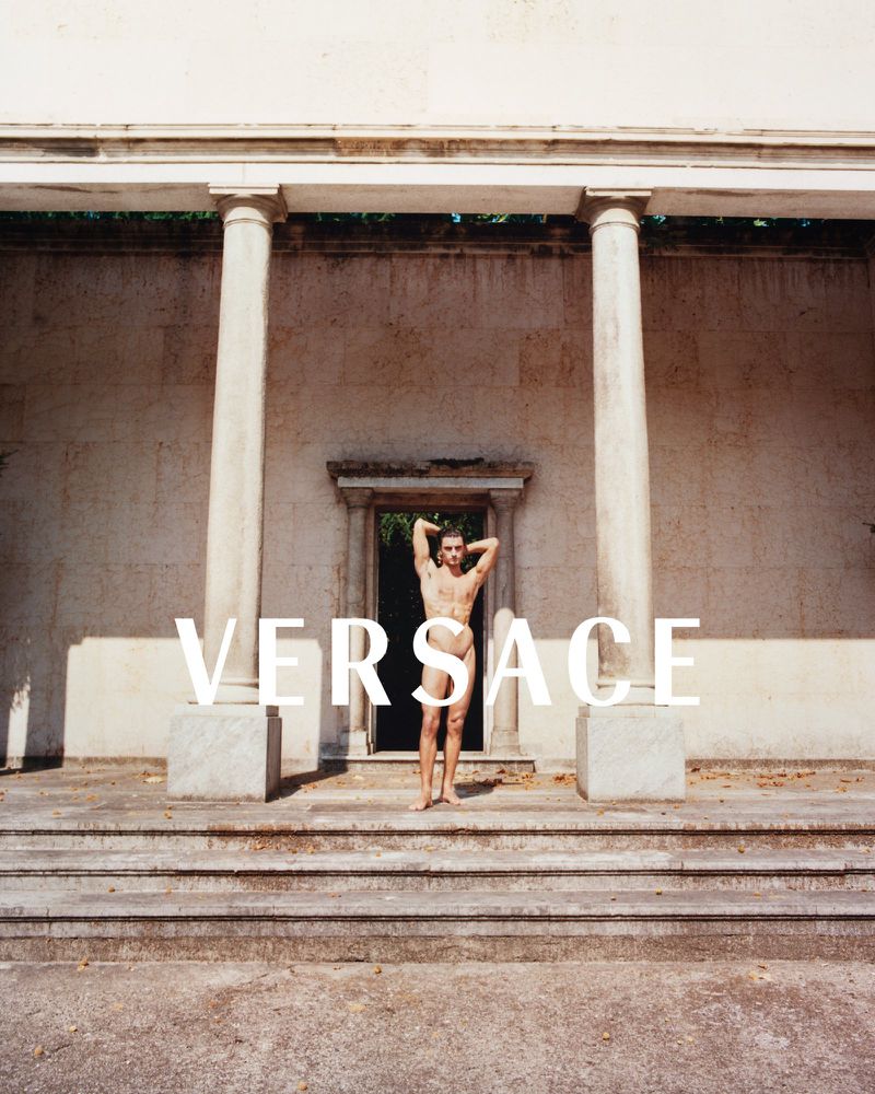 Nude adult male posing on steps in front of a building with columns and arched doorway, the brand VERSACE is superimposed across the image.