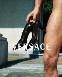 Close-up of a person holding a pair of black high heels with gold details, with Versace text overlaid on the image.