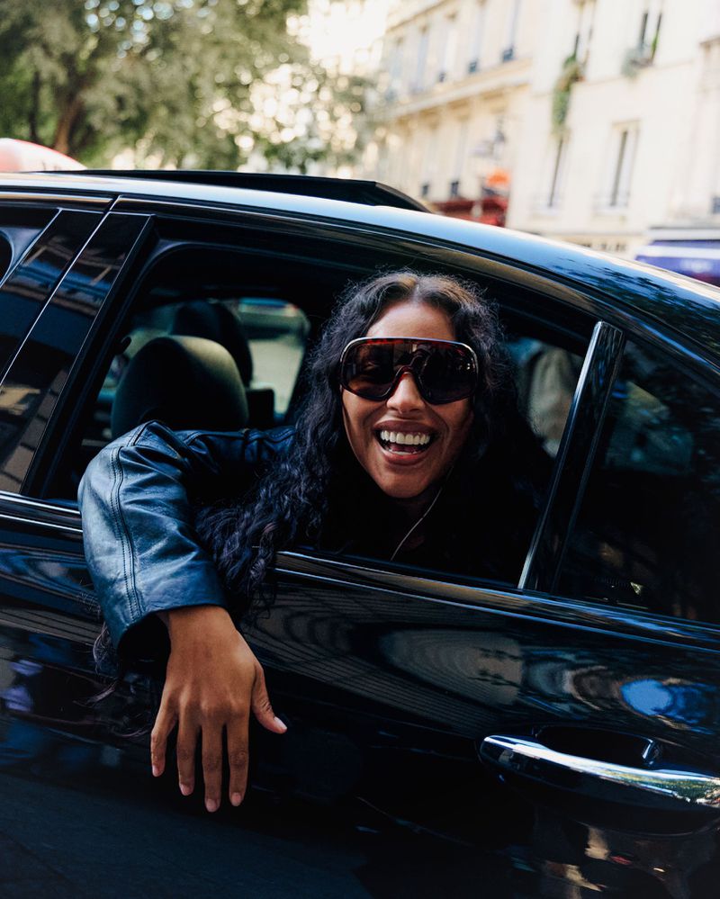 Smiling woman in sunglasses leans out of a black car in city. She wears a leather jacket and the urban setting is bright and sunny.