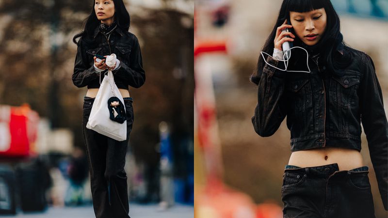 Street-style shot of an Asian woman with long dark hair wearing a cropped denim jacket and low-rise jeans, carrying a tote bag.