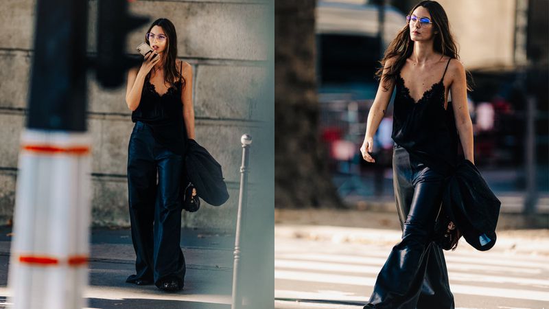 A woman walks on a crosswalk in a black camisole top and shiny, loose black pants. The woman has long brown hair and is holding a phone.
