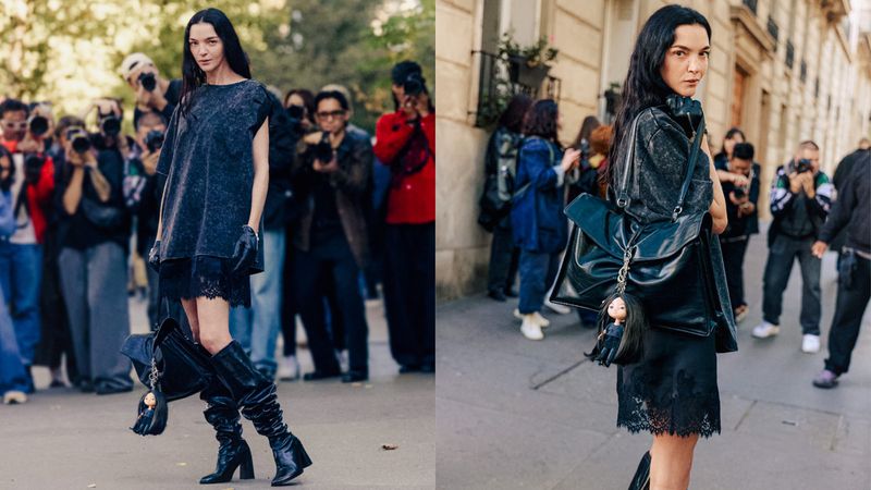 Fashionable adult female poses in a dark dress and knee-high boots on a city street with a crowd of photographers during the day.