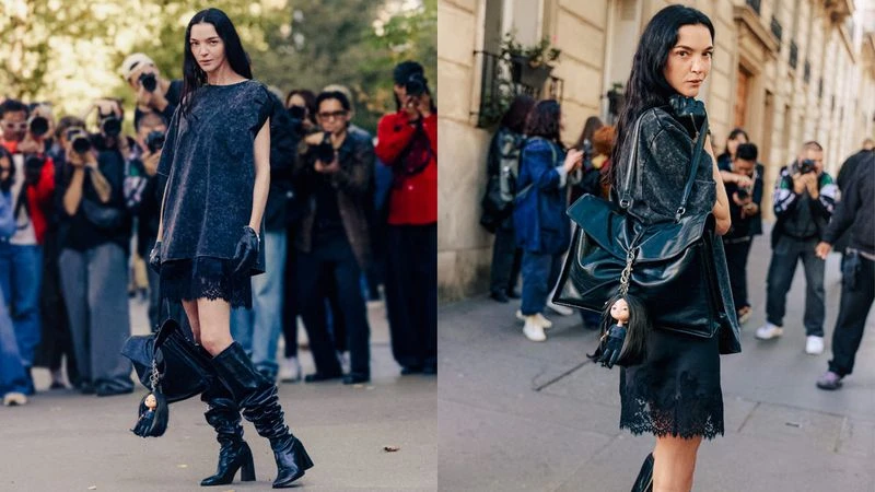 Fashionable adult female poses in a dark dress and knee-high boots on a city street with a crowd of photographers during the day.