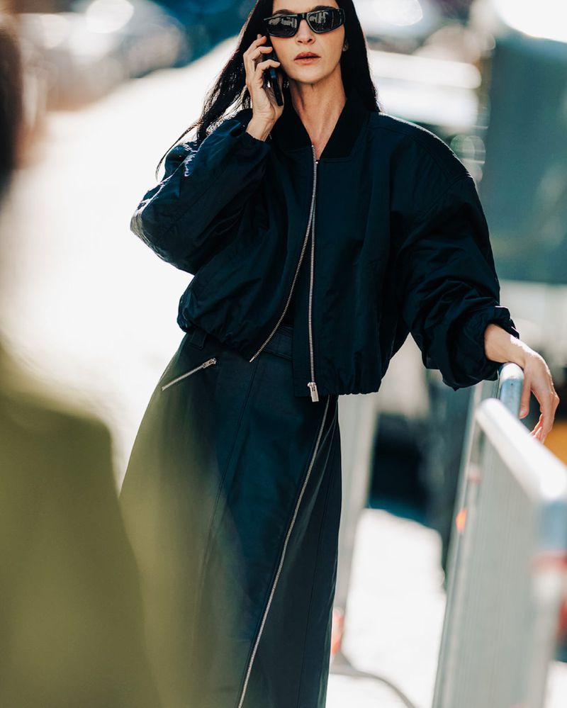 Brunette adult female in a navy bomber jacket and maxi skirt speaks on a mobile phone in an outdoor urban setting during daytime.