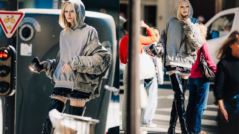 A slender woman with bleached hair walks on a city street wearing a grey hoodie, jacket, mini-skirt, and thigh-high boots with traffic nearby.